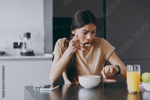 Young sad displeased disappointedhousewife woman 20s in t-shirt eat breakfast in morning look at smart watch check time hurry rush cooking food in light kitchen at home Healthy diet lifestyle concept.