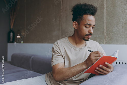 Pensive young poet student african american man 20s wearing beige t-shirt sit on grey sofa indoors apartment writing down poem in notebook diary, resting on weekends staying at home, enjoying hobby