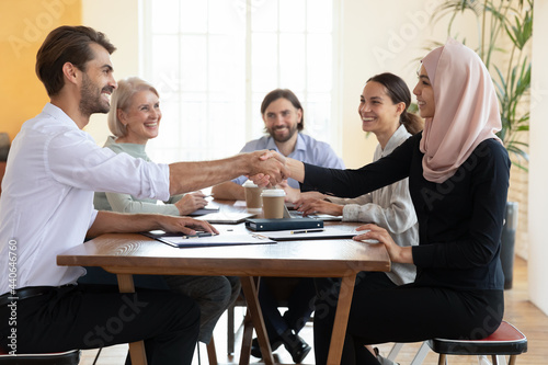 Diverse business partners handshaking, celebrating contract signing, sitting at table, successful group negotiations in boardroom, Asian muslim businesswoman wearing hijab shaking hand of businessman