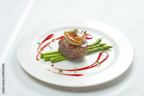 Portion of grilled mignon steak with foie gras and asparagus, berry sauce in a white plate. Isolated on grey background.