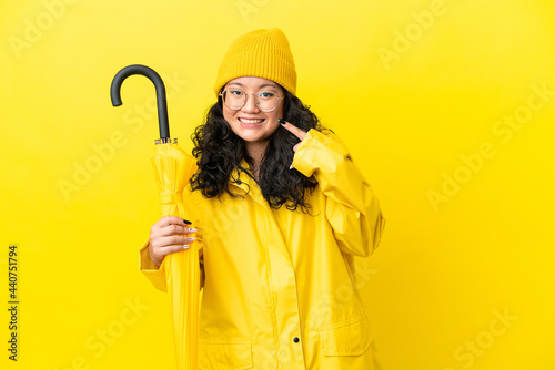 Asian woman with rainproof coat and umbrella isolated on yellow background giving a thumbs up gesture