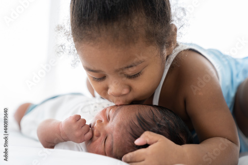 Cute African American little girl kissing on newborn baby cheek on white bed at home. Little girl takes care of infant baby with kindly