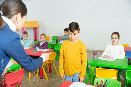 Upset boy listening reprimanding of strict female teacher during lesson in primary school