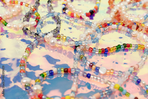 Closeup of necklaces and bracelets made from colorful beads and pearls on a pink holographic background.