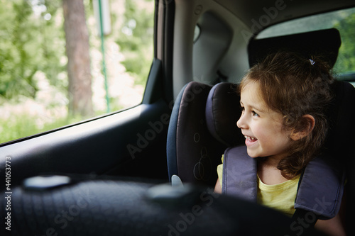 Adorable baby girl enjoys the travel by car, looks out of an opened window while sitting in a safety booster car seat