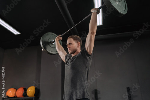 Portrait of muscular weightlifter doing an exercise with a barbell. Barbell above the head. Crossfit athlete having workout with barbell, doing military press, warming up. Dark gym background