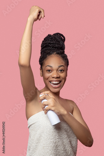 Dark-skinned young woman spraying deodorant on her body after shower