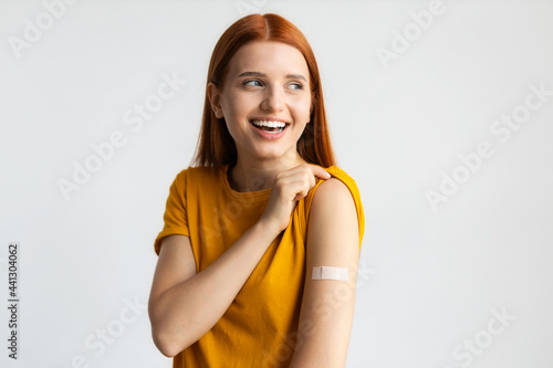 Covid-19 vaccinated caucasian smiling young woman showing arm with plaster, gray background