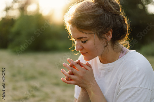 Woman closed her eyes, praying in a field during beautiful sunset. Hands folded in prayer concept for faith, spirituality and religion.