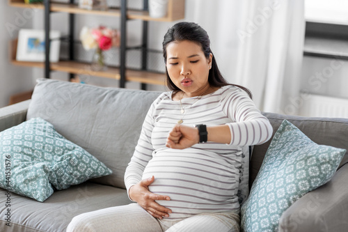 pregnancy, rest, people and expectation concept - pregnant asian woman with smart watch having labor contractions sitting on sofa at home and breathing