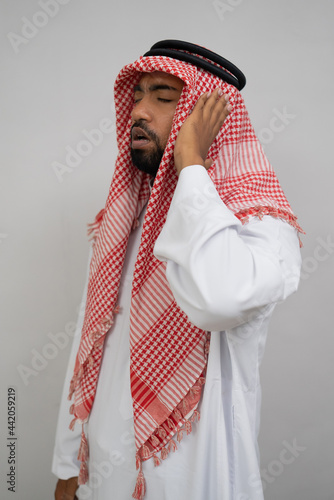 An Arab muezzin in a turban performs the call to prayer with one hand beside his ear on a plain background