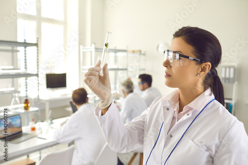 Side view young female scientist in coat, gloves and goggles looking at small green leaf inside test glass tube in her hand. Nature and science, using plants in cosmetology laboratory research concept