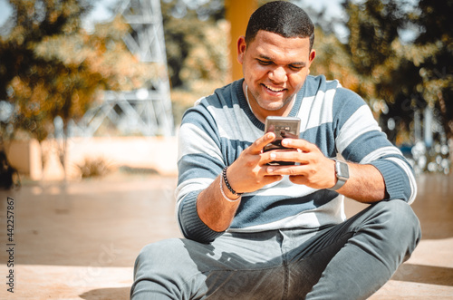 latin dominican man texting with his phone while smiling, sitting in the park