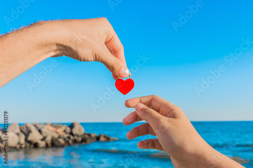 The man's hand holds a red heart next to the girl's hand against the sea coast background with a blue sky and a skyline. Sign and symbol of love and relationship concept