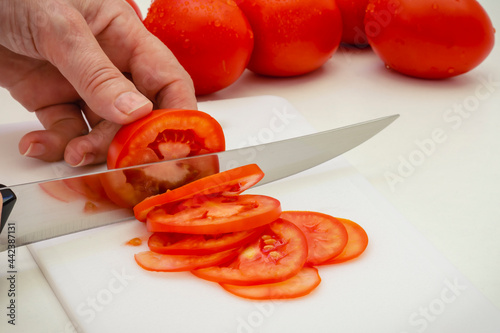 Manos con cuchillo cortando rodajas de tomate maduro sobre una mesa blanca