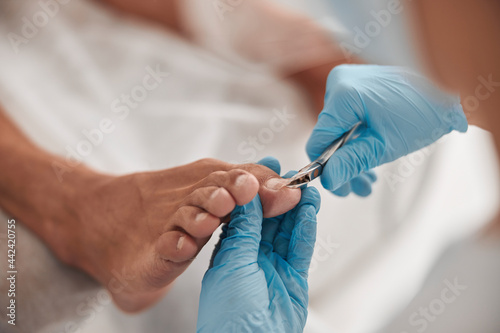 Master in protective gloves uses modern clipper to cut toenail of male client in salon