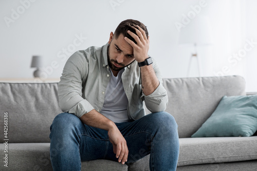 Unhappy man touching hair, sitting on sofa at home, thinking about problems