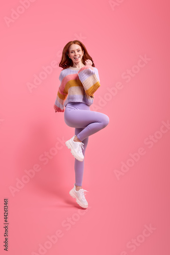 Attractive funny redhead lady jumping high raising hands first place winner wearing casual sweater, leggins isolated over bright pink color background. Beautiful lady rejoicing