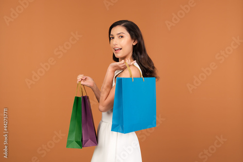 Half Asian-American woman in a cute white dress holding several colorful shopping bags in her hand. A Female happily shopping during sale season isolated on brown studio background.