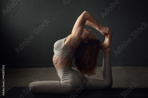 A slim beautifully lighted young woman is doing One-footed King Pigeon yoga pose ose (Eka Pada Rajakapotasana) in a dark room. Image with selective focus and toning