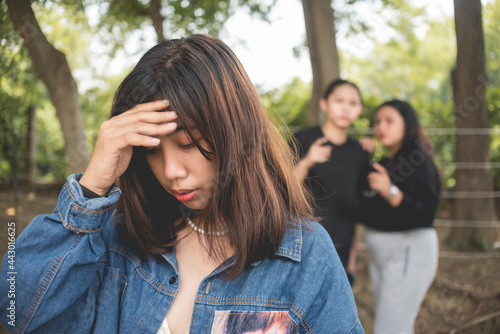 A depressed young woman holds her forehead in shame while 2 women talk behind her back. Teenage bullying or backstabbing.