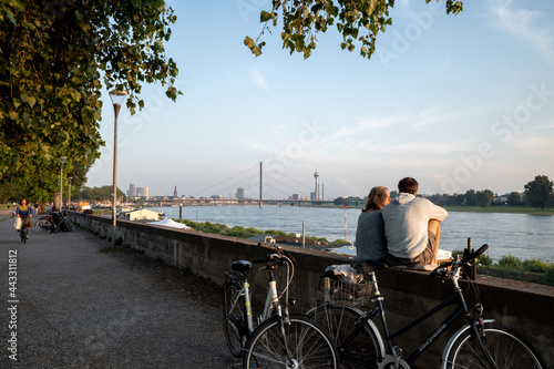 Outdoor sunny view, couple sit and chill out on balustrade at Rheinuferpromenade along riverside of Rhine river in Düsseldorf, Germany.