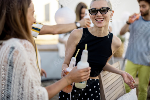 Young group of stylish people having fun at party outdoors, two female friends drinking cold drinks and talking at picnic on a roof top terrace