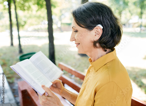 Mature woman with a hearing impairment uses a hearing aid in everyday life, reading a book in park, outdoor. Hearing solutions