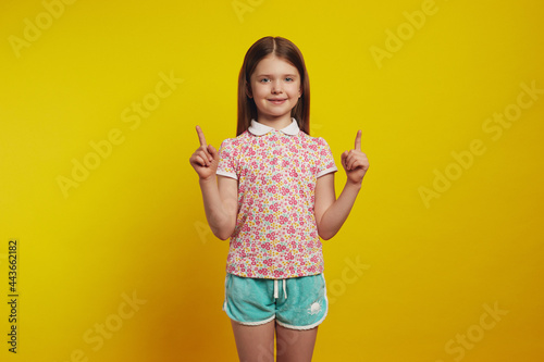 Friendly looking lovely girl kid shows promo with joy, points above with both index fingers, gives recommendation, wears stylish summer clothes