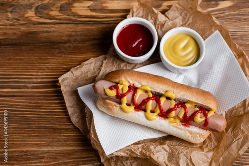 top view of hot dog near sauce bowls, paper napkin and crumpled kraft parchment on wooden table