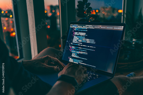 The code is on a laptop on a wooden table in front of the window in the dark with a view of the lights of the night city, color lighting in the room, home decor 