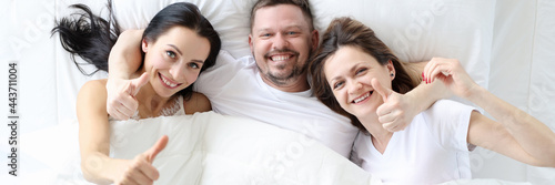 Two smiling women with man lie in bed and hold their thumbs up