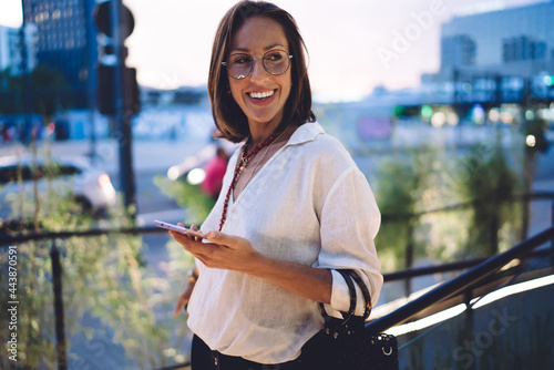 Cheerful beautiful woman in trendy wear and eyeglasses for vision protection laughing from funny social media content, pretty female blogger 30s holding mobile phone and rejoicing outdoors