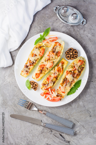 Ready-to-eat baked zucchini halves stuffed with cheese and tomato and basil leaves on a plate on the table. Vegetable menu, healthy food. Vertical and top view