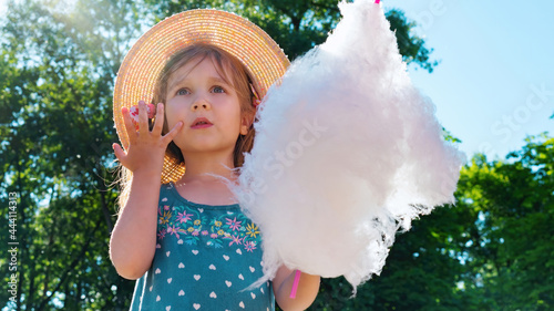 The girl looks in amazement and surprise at the amusement park. A child eats cotton candy on a sunny summer day and licks sticky fingers. Walk with children in the park on vacation.