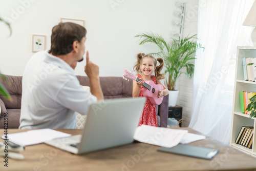 father works at home with laptop together with daughter. little girl wants to play guitar, noise and interfere with work. man shakes his finger at her and asks them to calm down remote work at home