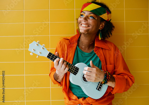 Fun rastafarian woman playing blue ukulele near yellow wall. Female street musician