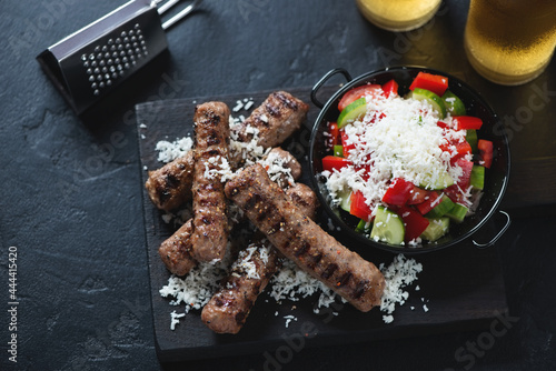 Grilled cevapi or serbian skinless beef sausages with grated bryndza and shopska salad, elevated view on a black stone background, horizontal shot