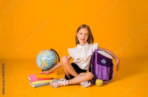 happy schoolgirl in uniforms is sitting with school supplies and a globe on a yellow background with a copy of the space