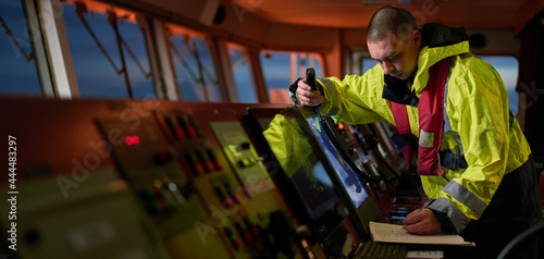 Navigator. pilot, captain as pat of ship crew performing daily duties with VHF radio, binoculars on board of modern ship with high quality navigation equipment on the bridge on sunrise.