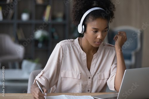 Close up focused African American woman in headphones taking notes, looking at laptop screen, sitting at desk, serious confident young female student watching webinar, listening to lecture, studying