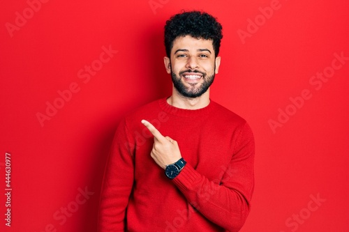 Young arab man with beard wearing casual red sweater cheerful with a smile of face pointing with hand and finger up to the side with happy and natural expression on face