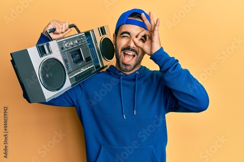 Young hispanic man holding boombox, listening to music smiling happy doing ok sign with hand on eye looking through fingers