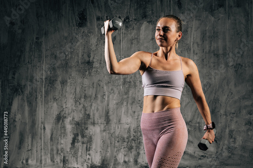 Middle shot portrait of muscular young athletic woman with perfect beautiful body wearing sportswear doing exercise with lifting dumbbells and looking away. Caucasian fitness female posing in studio.