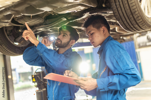 Two professional look technician inspecting car underbody and suspension system by using check list in moder car service shop. Automotive business or car repair concept. 