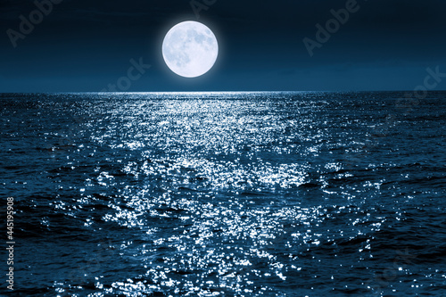 This large full blue moon rises brightly over the calm ocean creating sparkles across the waves 