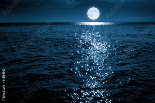 This large full blue moon rises brightly over the calm ocean creating sparkles across the waves