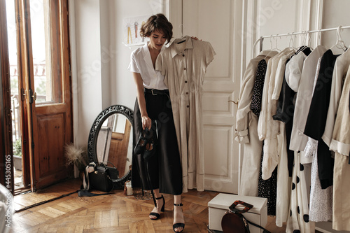 Stylish attractive short-haired woman in midi black skirt and white blouse poses in dressing room, holds trendy linen beige dress and shoes.
