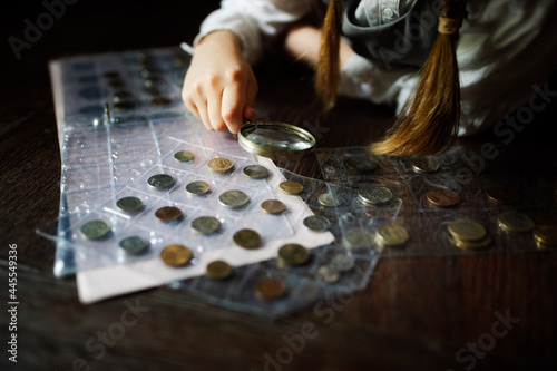 Cute cute european girl child in glasses with magnifying glass looks at coins, concept children and financial literacy, children and money, coin collecting and numismatics, dark style