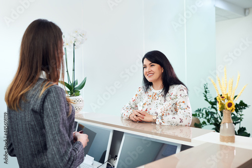 Mid adult woman with beautiful smile talking with young female receptionist, checking appointment in dentist clinic